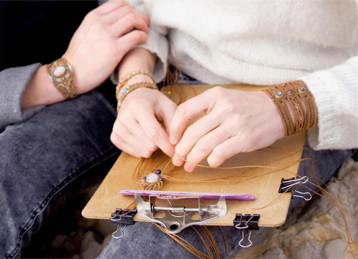 Mains tissant un bracelet en micro-macramé sur une planche de travail, poignets ornés de bracelets